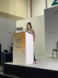 Woman standing on a podium at the conference IROS 2024 Abu Dhabi. She has black long hair put into a ponytail and glasses, wearing a beige loose blouse and black pants and pink and white sneakers. She is speaking into a microphone integrated in the podium and has a silver notebook open.