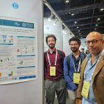 On the left: "Incremental Learning of Robotic Manipulation Tasks through Virtual Reality Demonstration" poster hanging on a white board with the number 6.13 above. On the right side of the poster, three men all in backpack (one man with curly dark brown hair and beard in wine red blouse and conference entry ticket on a white chainholder around his neck and grey pants with a brown belt, one man with brown straight hair and glasses and beard in a blue costume and blouse with the ticket on a white chainholder around his neck as well and the third man is bald with glasses and brown costume and light blue blouse) are standing side by side looking into the camera.
