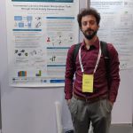 Right side in the picture: Man with dark brown curly hair and beard with a backpack on and in wine red blouse and grey pants with a brown belt on. He also has the conference entry card that is yellow around his neck on a white chainholder. In the background we can see a poster fixed to a white board and it is showcasing the paper on "Incremental Learning of Robotic Manipulation Tasks through Virtual Reality Demonstration". At the bottom right behind the man there is a backpack and another rolled up poster. Far behind we can see another poster hanging on a second white board.