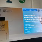 Man with curly dark brown hair and beard standing on a podium at a conference called IROS 2024 in Abu Dhabi. The stage is all white and there is a projection of a presentation: Learning Robotic Manipulation Tasks based on Incremental Demonstrations in a Virtual Environment, Logos of all participants, a deer in gold in the background (the deer is the main image of the conference).